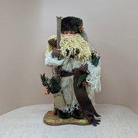 A tall, standing Father Christmas figure in a tan coat and brown fur hat. He has a long curly beard, holds a wooden staff, and carries a burlap sack with pine needles.