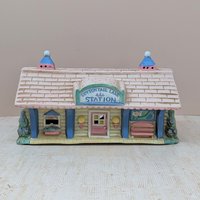 A vintage ceramic lighted house titled Cottontail Lane Station. It features a pink shingled roof with blue cupolas and a yellow clapboard exterior. The front includes a ticket window, a pink door, and a tiny bench under a "You Are Here" map.