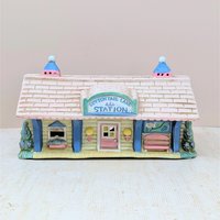 A vintage ceramic lighted house titled Cottontail Lane Station. It features a pink shingled roof with blue cupolas and a yellow clapboard exterior. The front includes a ticket window, a pink door, and a tiny bench under a "You Are Here" map.