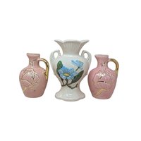 A three-piece vintage pink ceramic vase lot. The centerpiece is a 6.5-inch Hull Magnolia vase with blue flowers and double handles. It is flanked by a pair of 5-inch pink pitcher bud vases featuring embossed leaf designs and gold-clad handles.