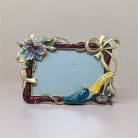 A horizontal photo frame with a deep red enamel border. Ornate gold-tone decorations include a crystal-studded purple hibiscus-style flower on the top left and a fashionable stiletto shoe on the bottom right.