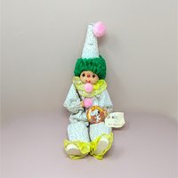 A playful clown doll sitting upright, wearing a white outfit with tiny colorful dots and yellow ruffles. It has green curly yarn hair and three pink pom-poms down its front, holding a small paint brush.