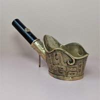 A traditional brass coal iron with a deep cup-shaped base and a black wooden handle. The exterior of the brass is decorated with raised scrollwork and ancient-style markings.