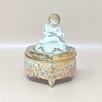 A vintage footed trinket box with a highly detailed gold-toned floral base and a white bisque-style lid featuring a cherub playing a stringed instrument.