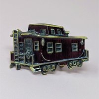 A vintage 1976 belt buckle shaped like a train caboose with deep red enamel and brass-toned metal accents on a neutral background.