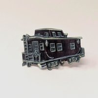 A vintage 1976 belt buckle shaped like a train caboose with deep red enamel and brass-toned metal accents on a neutral background.
