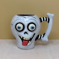 A whimsical white ceramic skull mug with large bulging eyes, a protruding red tongue, and a black-and-white striped bone handle against a neutral background.