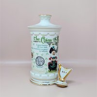 A tall white porcelain decanter featuring colorful Irish coin graphics, a leprechaun illustration, and a gold-trimmed harp stopper against a neutral background.