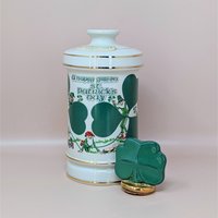 A tall white porcelain decanter featuring green "A Happy Green St. Patrick’s Day" typography, leprechaun illustrations, and a gold-trimmed shamrock stopper against a neutral background.