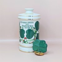 A tall white porcelain decanter featuring green "A Happy Green St. Patrick’s Day" typography, leprechaun illustrations, and a gold-trimmed shamrock stopper against a neutral background.