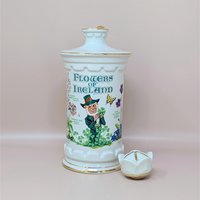 A white porcelain decanter featuring botanical "Flowers of Ireland" illustrations, a leprechaun figure, and a gold-trimmed flower-shaped stopper against a neutral background.