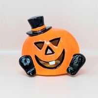 A glossy orange ceramic pumpkin figurine wearing a black top hat and featuring a single white tooth against a neutral background.