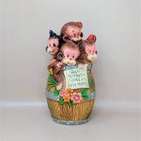 A whimsical hand-painted ceramic bank featuring four brown monkeys peering out of a wooden-style barrel with a motivational savings scroll against a neutral background.