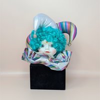 A porcelain jester doll head with teal hair and a rainbow-striped hat emerging from a black velvet cube against a neutral background.