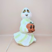 A large white ceramic ghost figurine with iridescent green highlights holding an orange jack o' lantern, featuring large expressive eyes and a wide smile.