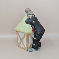 A bisque porcelain figurine of a sad clown in tattered black clothes leaning against a pale yellow street lamp against a neutral background.