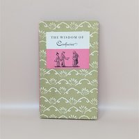 A slim hardcover book with a sage green and cream floral pattern, featuring a central pink and white label with the title The Wisdom of Confucius and a small illustration of three figures against a neutral background.