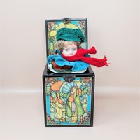 A black wooden music box with an open lid, revealing a porcelain boy doll with a green hat and red scarf. The box features colorful illustrations of Bob Cratchit and Tiny Tim.