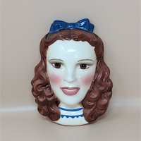A vintage hand-painted ceramic wall mask of Dorothy from The Wizard of Oz, featuring her iconic pigtails and a bright blue hair bow.