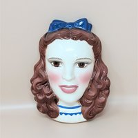 A vintage hand-painted ceramic wall mask of Dorothy from The Wizard of Oz, featuring her iconic pigtails and a bright blue hair bow.