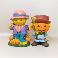 A pair of vintage hand-painted ceramic figurines featuring a friendly scarecrow standing on pumpkins and a jack o' lantern man holding a broom.