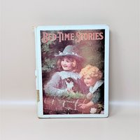 An antique 1914 hardcover book titled Bed-Time Stories featuring a front cover illustration of two children with a kitten and an interior plate of children playing around a snowman