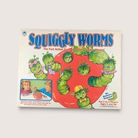 Squiggly Worms board game box cover featuring illustrated cartoon green worms popping out of a large red apple.