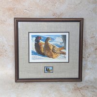 A 1987 Minnesota Pheasant Habitat Stamp Print by Brian Jarvi on a tan textured background. The framed art features a signed and numbered print of a pheasant pair in the snow, a matching stamp below, an oatmeal linen mat, and a dark wood frame.