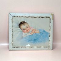 A 1940s vintage Charlotte Becker framed nursery print on a soft pink background. The sweet lithograph depicts a rosy-cheeked baby tucked under a blue blanket, housed in a distressed white decorative wooden frame with chipped paint accents.