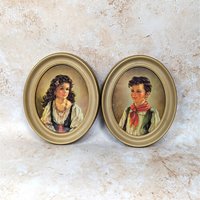 A pair of 1950s vintage Ann Allaben framed oval prints on a tan textured background. The matching set features the portraits of a young Spanish girl named Elena and a boy named Chico, both dressed in traditional mid-century clothing inside gold oval frames.