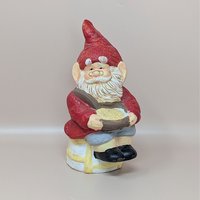 A vintage ceramic gnome figurine with a tall red hat, white beard, and glasses, sitting on a stool and holding a golden bowl.