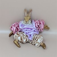 A vintage 1990s rabbit figurine in a seated pose, wearing voluminous ruffled satin garments in pastel lavender and rose pink with a white lace collar and small golden shoes.Measurements: Approximately 6.5" Height  Decade: 1990s  Style: Whimsical Pierrot  Material: Satin, Resin, Lace  Condition: Very Good Vintage Condition  Features: Hand Painted Details, Ruffled Satin Clothing, Seated Pose