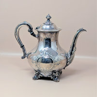 A vintage silver plated teapot with ornate hand chased floral scrollwork and a decorative finial on the lid, standing on four scrolled feet