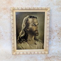 A 1950s vintage Warner Sallman Head of Christ framed lithograph on a tan textured background. The nineteen inch portrait of Jesus is housed behind glass in a distressed, cream painted notched wooden frame showing natural age marks.