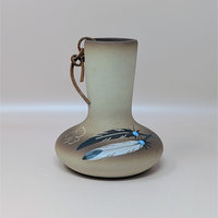 A hand painted tan pottery vase by Betty Selby featuring black and white feather motifs with turquoise accents and a leather strap tied around the neck.