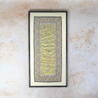 An antique Chinese silk embroidery sleeve band framed wall hanging on a tan textured background. The long vertical gold silk panel features intricate hand stitched figures and symbols inside a decorative patterned textile border and a dark wood faux bamboo frame.