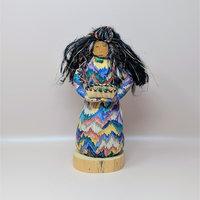 A handmade folk art doll by Patty Kune, featuring a figure in a vibrant, multi-colored zigzag patterned dress. The doll has long black fiber hair and holds a small woven basket, standing on a circular wooden base against a neutral background.