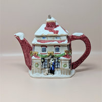 A ceramic teapot shaped like a snow-covered Dickensian cottage with a brick chimney and festive garlands. A small figure in a top coat stands at the front door with a dog. The spout and handle have a red brick texture.