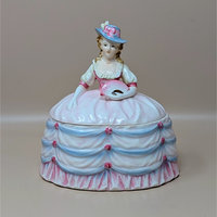 A vintage ceramic powder box shaped like a Victorian lady in a billowing pink and blue dress. She wears a wide-brimmed hat and holds a pink fan. The base of the skirt acts as the lid for the trinket dish.