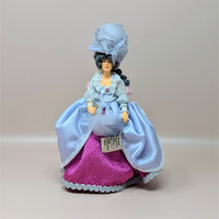A vintage Peggy Nisbet portrait doll inspired by a Gainsborough painting. She wears a lavish lavender satin overskirt with a bright pink textured underskirt, a ruffled bodice, and a large feathered blue hat.