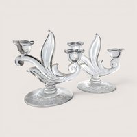 A pair of elegant clear glass candelabras with curved leaf details and flower-shaped cups, centered on a beige background.