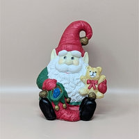 A vintage hand-painted ceramic music box shaped like a seated Santa elf. He has a long white beard, a pointed red hat with a bell, and is holding a small teddy bear and a green toy sack.
