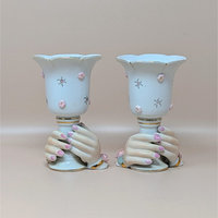 A pair of vintage ceramic candle holders featuring a pair of hands with pink fingernails clasping the base of a white cup. The cups are decorated with small pink 3D flowers and gold-rimmed scalloped edges.