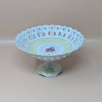 A vintage porcelain pedestal bowl featuring a reticulated lace-cut rim and a central Fragonard-style illustration of a courting couple. The bowl has a pale yellow border and delicate gold-gilded accents throughout.