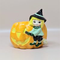 A vintage ceramic Halloween planter featuring a cute blonde witch riding on the side of a large orange jack-o'-lantern. The witch wears a green tunic and a black pointed hat with a mint green stripe.