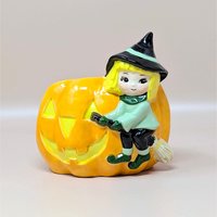 A vintage ceramic Halloween planter featuring a cute blonde witch riding on the side of a large orange jack-o'-lantern. The witch wears a green tunic and a black pointed hat with a mint green stripe.