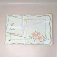 A vintage 8-piece Bucilla table linen set featuring four placemats and four matching napkins with colorful cross-stitch floral designs. The cream-colored linens are tied together with a white ribbon.