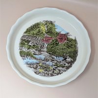 A vintage Johnson Brothers ceramic pie plate featuring a classic transferware scene from The Friendly Village pattern. The plate has a scalloped rim and depicts a red barn near a stream with water lilies.