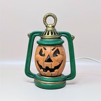 A vintage ceramic jack-o'-lantern light shaped like an old-fashioned lantern. The pumpkin is orange with a carved grin, set inside a green frame with gold detailing. An electrical cord is attached for illumination.