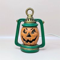 A vintage ceramic jack-o'-lantern light shaped like an old-fashioned lantern. The pumpkin is orange with a carved grin, set inside a green frame with gold detailing. An electrical cord is attached for illumination.